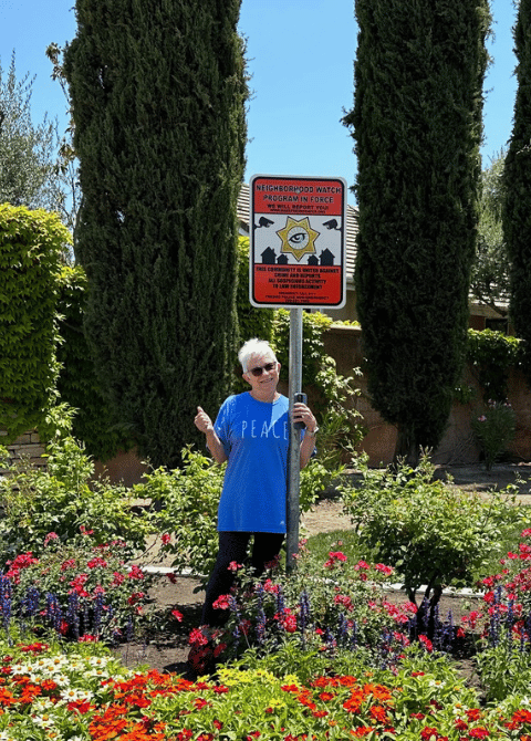 Sue Kendall stands next to a neighborhood watch sign in a flower garden.